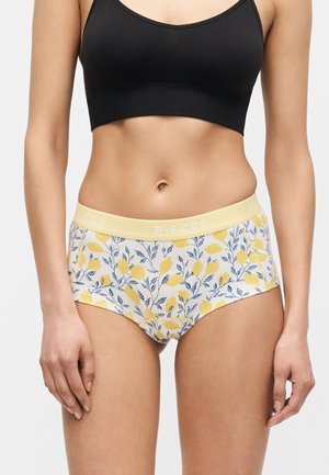 Kvinne ikledd svart cropped tanktop og hvite høyhøye undertøy med gul sitron- og blå bladmønster, står mot ensfarget bakgrunn.