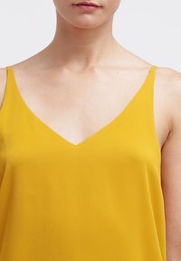 Gul, ärmlös topp med V-ringning i ett mjukt tyg. Har tunna axelremmar och en löst sittande passform.