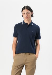 Jūras spēku polo krekls ar baltām svītrām apkaklītes un piedurkņu malās, īsām piedurknēm un mazu logotipu uz krūškurvja. Saskaņots ar khaki biksēm.