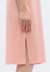 Φόρεμα μέχρι το γόνατο σε απαλό ροζ ύφασμα, με πλαϊνή σχισμή και ίσιο τελείωμα. Η ραφή προσθέτει διακριτική δομή.