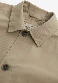 Camicia beige con colletto, caratterizzata da una chiusura con bottoni e una texture sottile. L'etichetta indica la taglia M; realizzata in un tessuto liscio.