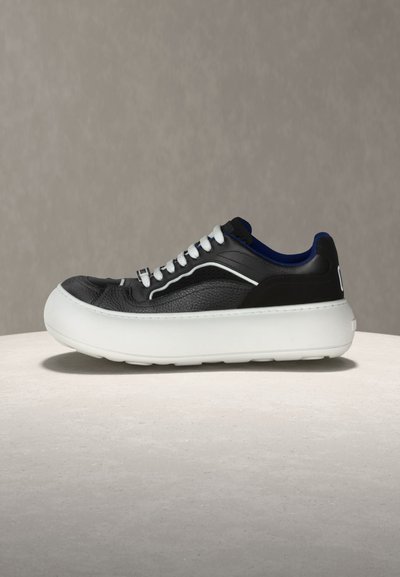 Juodi žemo profilio sportbačiai su stora balta platformos padu, baltais batraiščiais ir mėlynu vidiniu pamušalu, padėti ant šviesiai pilkos dangos prie tamsiai rudos fono.