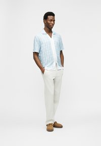 Νέος άνδρας που στέκεται με τα χέρια στις τσέπες, φοράει ανοιχτό μπλε πουκάμισο με κοντά μανίκια και σχέδια, λευκό παντελόνι και καφέ παπούτσια, πάνω σε λευκό φόντο.