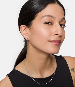 Orecchini d'argento con un design lineare e pendenti; collana a catena d'argento con piccoli dettagli di perline; indossata sopra una maglia nera.