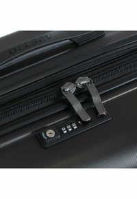 Black hard-shell suitcase with a ridged texture, featuring dual metallic zippers and a combination lock with a keyhole. Brand name "Delsey" visible.