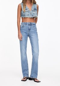 Veste courte en denim avec col en V, dotée de poches et d'un ourlet effiloché ; associée à un jean droit taille haute de couleur bleu clair. Sandales noires ouvertes à l'avant.