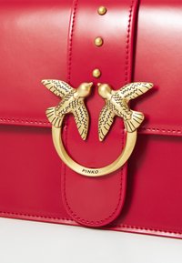 Červená kožená kabelka s zlatu kruhovou sponou a dvoma ozdobami vo forme vtákov, zdobená gravírovanými vzormi. Sú viditeľné aj vyšívané detaily.
