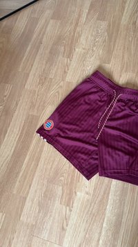 Maroon athletic shorts featuring vertical stripes, an elastic waistband with a drawstring, and a circular logo on the left hem.