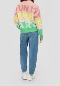 Kvinna i en flerfärgad tröja med stor text, blå jeans, vita strumpor och klumpiga vita sneakers, som står mot en enfärgad bakgrund.