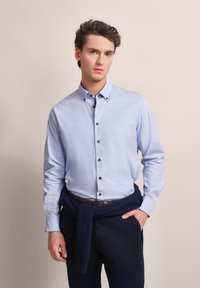 Helles blaues Hemd mit braunen Knöpfen, kombiniert mit dunkelblauen Hosen. Ein dunkelblauer Pullover ist um die Taille gebunden. Glatte Stoffoberfläche.