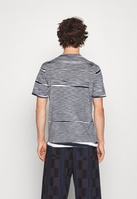 Modelo masculino visto desde atrás, vestido con una camisa de manga corta a rayas y pantalones oscuros con patrón, contra un fondo blanco liso.
