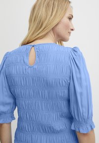 Lys blå bluse med smocket tekstur, puffede ærmer og en nøglehulslukning bagpå med en lille knapdetalje. Let stof.