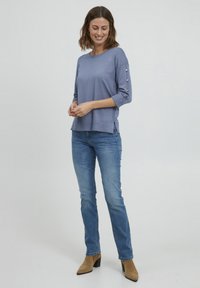 Lila gestrickter Oberteil mit dreiviertel Ärmeln und Knopfakzenten an den Ärmeln, kombiniert mit blauen Jeans und beigen, hochhackigen Stiefeletten.