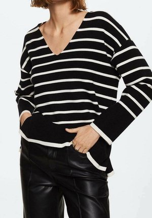 Person iført en sort og hvid vandret stribet V-hals sweater og sorte læderbukser, hænderne i lommerne, beskåret ved nakken og hofterne.