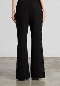 Pantalones flare negros con paneles de encaje en los laterales. Tejido suave, cintura alta, ligero brillo en el encaje, sin bolsillos visibles, acentos metálicos en el dobladillo.