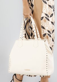 Borsa a tracolla color crema con dettagli a borchie, forma strutturata, doppi manici e superficie liscia. Presenta un logo sul davanti.