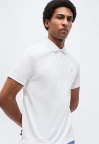 Νεαρός άνδρας με κοντά, σγουρά μαλλιά, φορώντας απλό λευκό πόλο με κοντά μανίκια και ένα μικρό λογότυπο στο στήθος, που στέκεται μπροστά σε λευκό φόντο.