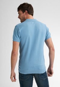 Světle modrá polokošile s krátkým rukávem a texturovaným vzorem, s žebrovaným límcem a rovnými okraji. Nosená s tmavě modrými džínami.