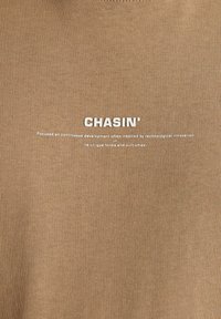 Rudas raukšlėtas medvilninės marškinėlių su balta logotipu "CHASIN'" centre ant krūtinės ir papildomu tekstu po juo.