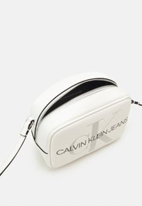 Biała torebka crossbody z sztucznej skóry z zamkiem błyskawicznym, z wyraźnym logo "CALVIN KLEIN JEANS" w kolorze szarym. Teksturowana powierzchnia, regulowany pasek.