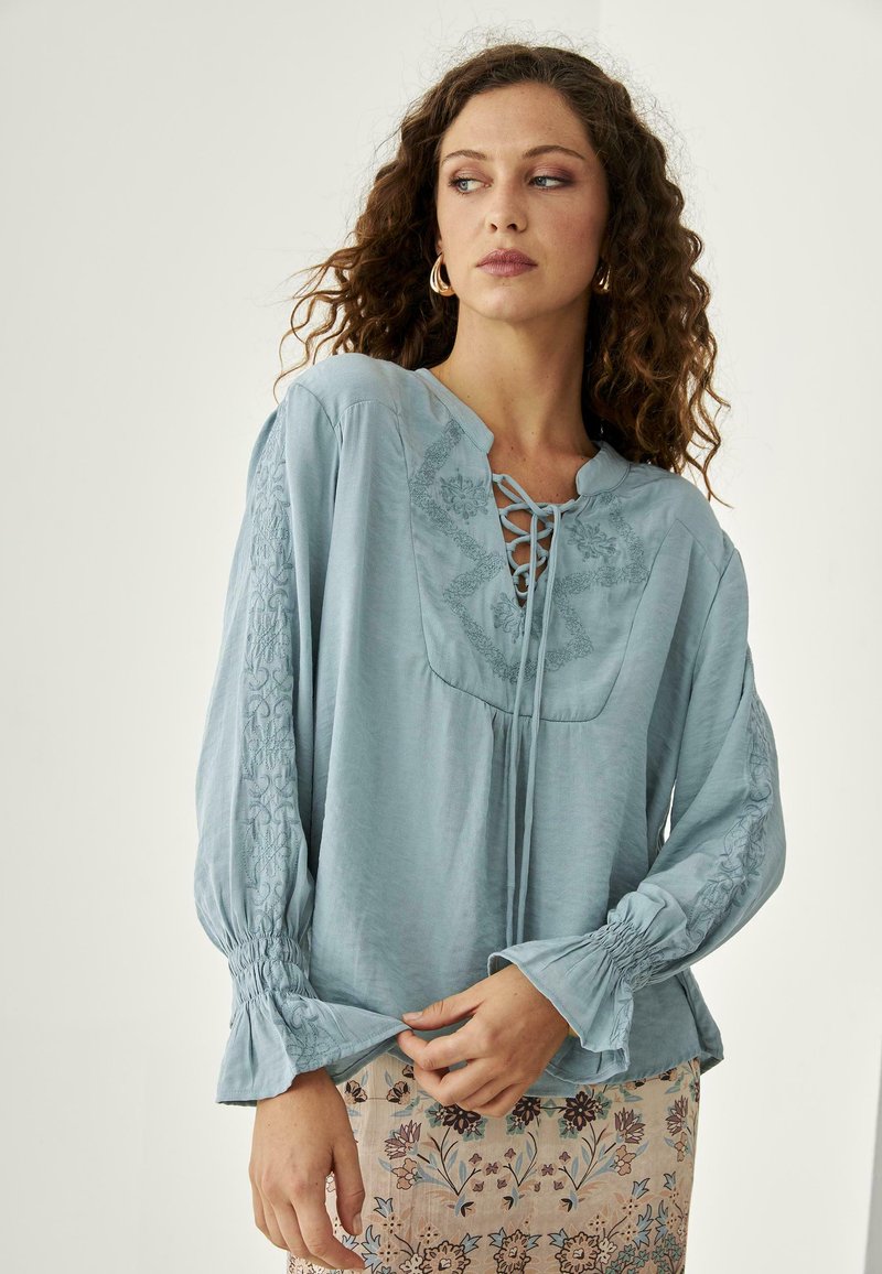 Mujer con cabello rizado que lleva una blusa bordada de color azul claro con escote de cordones y una falda de patrón floral, mirando de lado.