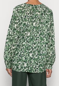 Personne portant un blouse à manches longues avec un motif floral à feuilles vertes et blanches, associée à un pantalon vert foncé, vue de dos.