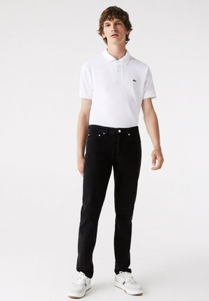 Polo shirt blanche à manches courtes avec col et logo vert. Jean noir slim avec cinq poches. Baskets blanches avec des accents gris.