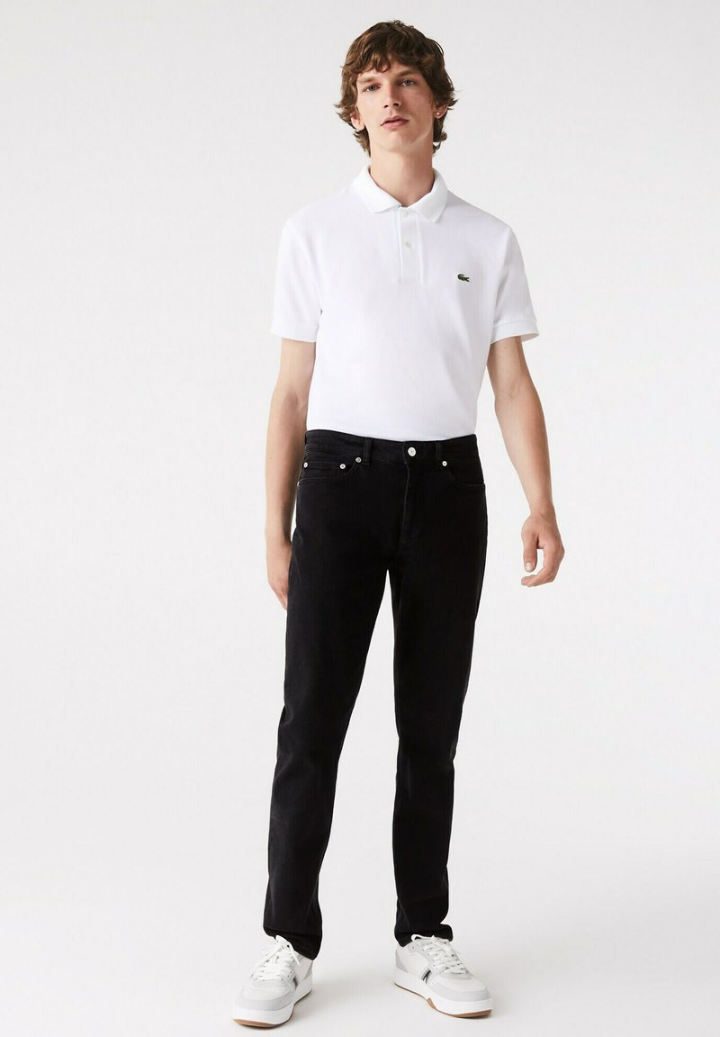 Weißes Poloshirt mit kurzen Ärmeln und Kragen sowie grünem Logo. Schwarze Jeans mit Slim-Fit und fünf Taschen. Weiße Sneakers mit grauen Akzenten.