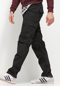 Pantalons cargo noirs avec plusieurs poches, coupe cintrée et texture lisse, associés à des chaussures de sport grises avec des rayures noires.