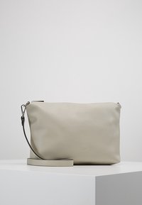 Pochette rectangulaire en cuir beige avec sangle de poignet amovible, fermeture éclair argentée et logo embossé discret, présentée sur un socle blanc.