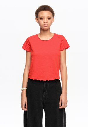 Joven con una blusa roja de manga corta y bajo con volantes, y pantalones negros de cintura alta, de pie frente a un fondo blanco liso.