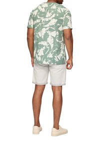 Grünes T-Shirt mit kurzen Ärmeln und weißem Blättermuster, kombiniert mit beigen Shorts und hellen Turnschuhen. Glatte Stoffqualität und lässige Passform.