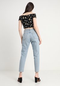 Černý květinový crop top s designem bez ramínek, v kombinaci se světle modrými džínami s vysokým pasem. Modelka má na sobě černé sandály na podpatku.