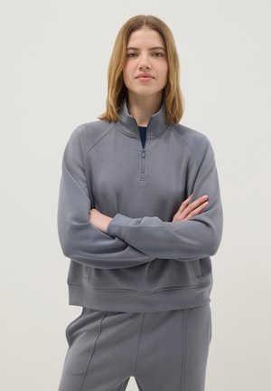 Jeune femme bras croisés portant un sweat-shirt gris à demi-fermeture éclair et un pantalon assorti, sur un fond clair uni.