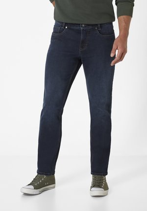 Dark blue jeans with a slim fit, featuring five pockets, contrasting stitching, and a button closure. Paired with green canvas sneakers.
