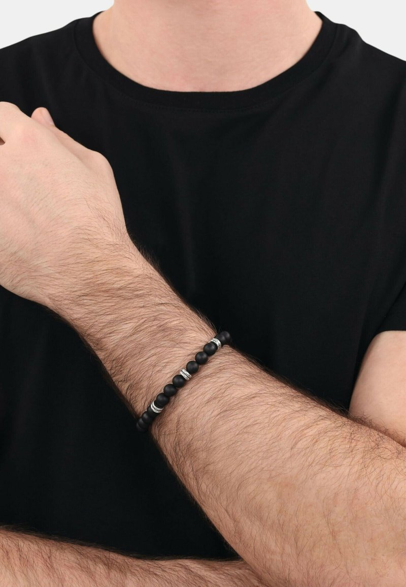 Bracciale nero con perline che presentano una combinazione di perline nere opache e lucide, decorate con perline d'accento argentate, indossato su un polso con peli visibili.