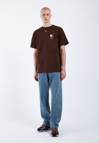 Camiseta de algodón marrón con un logo de corazón blanco, combinada con vaqueros de corte claro y zapatillas marrones con acentos en negro y blanco.