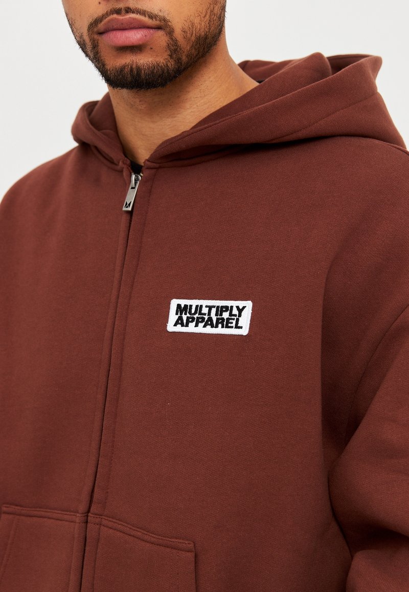 Homme portant un sweat à capuche marron zippé, capuche abaissée, avec un patch blanc sur la poitrine portant l'inscription « MULTIPLY APPAREL ».