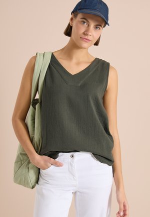 Femme portant un haut sans manches vert foncé, pantalon blanc, casquette bleue, portant un sac matelassé vert clair sur l'épaule, debout sur fond beige.