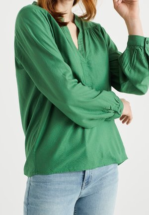 Osoba ubrana w zieloną bluzkę z długim rękawem z plisowanymi mankietami oraz jasnoniebieskie dżinsy, poprawiająca rękaw na białym tle.