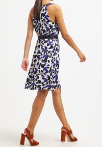 Robe fleurie blanche, bleue et noire, avec une taille cintrée et un dos ouvert. Assortie à des sandales à talons marron.