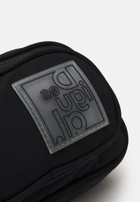 Černá polstrovaná taška s texturovanou, průhlednou nášivkou loga. Zipper a švy jsou viditelné podél okrajů.