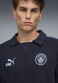 Maglione blu navy con colletto, con logo del Manchester City e scritta "KING" stampata in bianco. Logo Puma sul lato sinistro del petto. Tessuto strutturato.