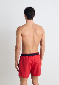 Shorts de bain rouges avec une taille élastique noire arborant le logo "HUGO". Tissu lisse et coupe décontractée, vus de l'arrière.