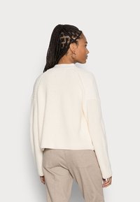 Suéter tricotado em tom creme, com um design cropped, textura canelada e ombros caídos. Apresenta um decote alto e mangas largas.