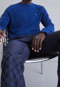 Blå stickad tröja med rund hals och liten logotyp på bröstet, tillsammans med mörka mönstrade byxor med ett upprepande mönster.