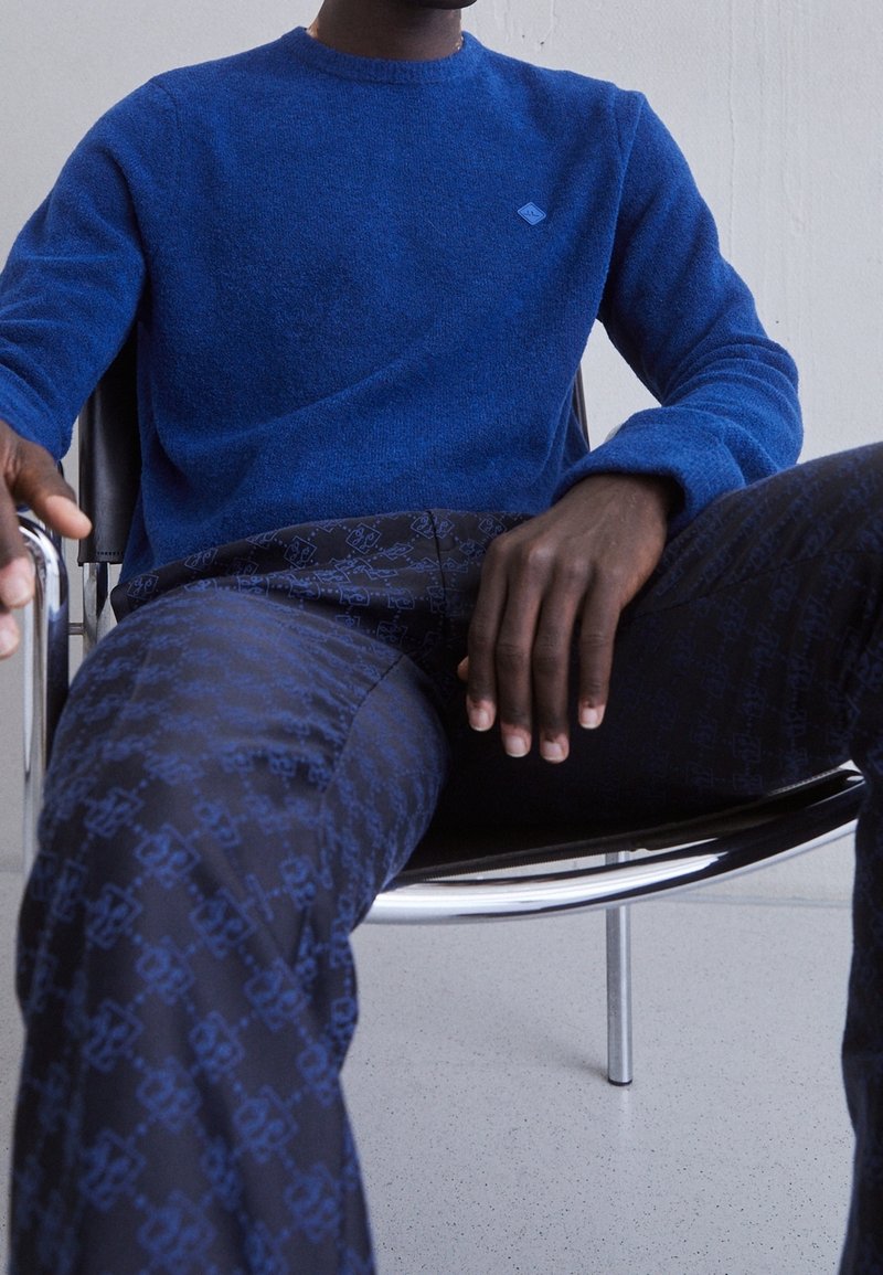 Blå stickad tröja med rund hals och liten logotyp på bröstet, tillsammans med mörka mönstrade byxor med ett upprepande mönster.