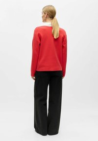 Sarkana džemperis ar apaļu kaklu un nolaižamām pleciem, kombinēts ar melnām platām biksēm. Gluda tekstūra ar ikdienišķu piegriezumu.