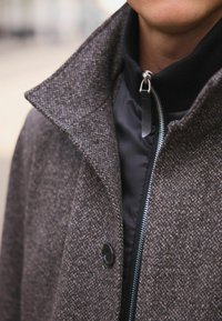 Brun tweedkappa med stor krage, utrustad med ett svart innerfoder, framdragkedja och detalj med knappstängning.