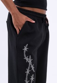Zwarte sweatpants met een barbed wire ontwerp in witte borduursel langs de linkerkant, voorzien van een elastische tailleband en zijzakken.